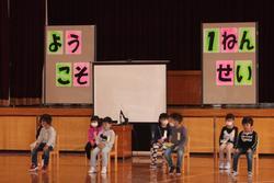 「ようこそ1ねんせい」と書かれたパネルの前で、1年生の児童が間隔をあけて座っている写真