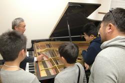 グランドピアノを弾いている初老の男性の周りに、参加者たちが集まっている写真