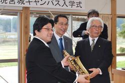 スーツの男性たちが、金色の管楽器を手渡している写真