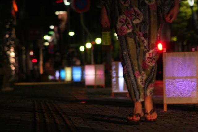 夜、路上に並んでいるカラフルな灯篭の横を、浴衣姿の女性が歩いている写真