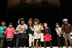 舞台の上で、子供たちと大人の女性が笑顔で立っている写真
