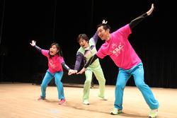 緑色の衣装を着た人と、ピンクのシャツを着た人たちが、手を一か所に合わせて踊っている写真