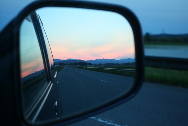 車のサイドミラーに、真っすぐな車道と夕焼けが見える写真