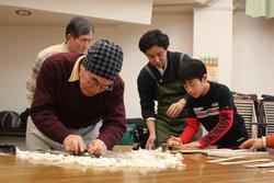 机の上で、初老の男性や男の子が楽器を制作している写真