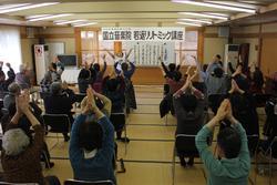 畳の部屋に集まった参加者たちが、両手を合わせて頭の上にあげる体操をしている写真