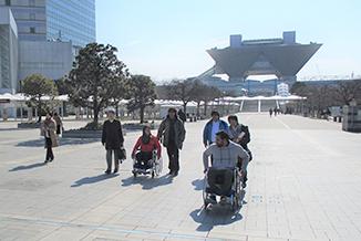 屋外で東京ビッグサイトを背景に歩く人達と車椅子の人たちの写真