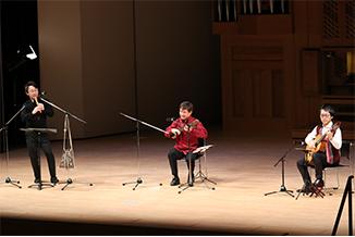 ホールの舞台上で楽器を演奏する男性三人の写真