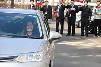 旗を持った職員たちを背に、笑顔の選手を乗せた車が走っていく写真