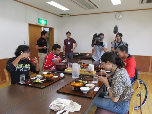 机に置かれた昼食を食べる男女とその様子をテレビ取材する人たちの写真