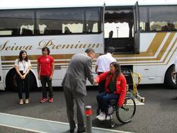 屋外でバスの前で握手をする背広の男性と車椅子の女性の写真