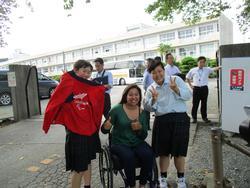 高校の屋外で撮られた車椅子の女性と高校生たちが横に並んで写った記念写真