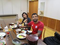 昼食が並んだ長机に向かい、食事をしながら笑顔でカメラを見ている選手たちの写真