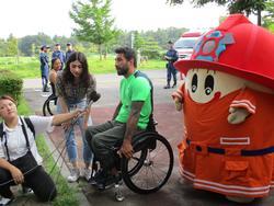 屋外で取材に応じる車椅子の男性と女性とマスコットキャラクターの写真