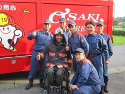 消防車の前で並んで映る車椅子の男性と救急隊員の記念写真