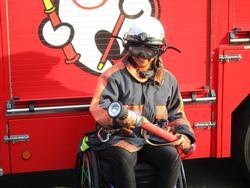屋外で消防車の前で消防士のユニフォームを着てポーズをとる車椅子の男性の写真