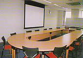 壁際にスクリーンが掛かり、その前に丸い形の長机が置かれている会議室の写真