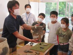 土器を持って見せている学芸員の男性と、それを眺めている生徒たちと先生の写真