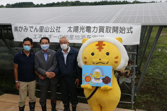 マスコットキャラクターがイラストを手に持ち、その隣で3名の男性が並んで記念撮影をしている写真